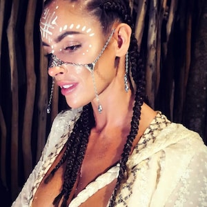 May include: A woman with dark braids and white face paint wears a silver face chain and earrings. She is wearing a cream-colored top with gold embellishments. The background is a dark, wooden texture.
