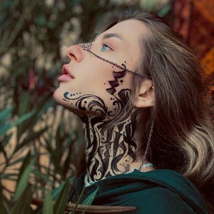 May include: A woman with long brown hair wearing a green hooded shirt and a silver chain across her face. The chain has a decorative design and is attached to a nose ring. The woman's face is painted with black ink in a decorative pattern.