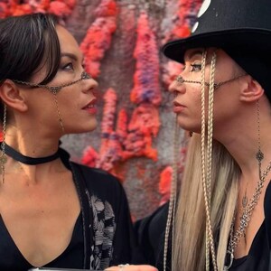 May include: Two people wearing black clothing and elaborate chain nose and face jewelry. The person on the right is wearing a black top hat with white polka dots. The person on the left is holding a black book with the word "witch" printed in white on the cover.