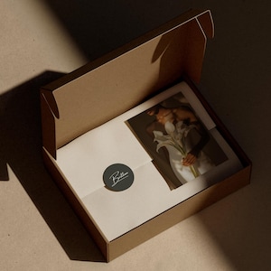 Op de afbeelding: Een open bruine kartonnen verzenddoos met daarin een witte envelop verzegeld met een donkergrijze ronde sticker met de witte tekst Britten, naast een rechthoekige print waarop een persoon met een boeket witte lelies is afgebeeld.