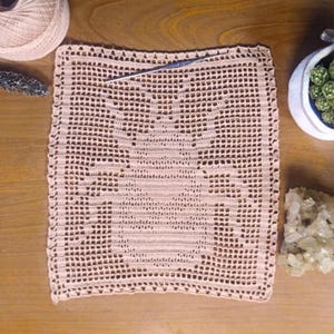 May include: A handmade, square, peach-colored crochet doily with a detailed openwork design. A crochet hook rests on the doily, with a ball of yarn and a small container of succulents nearby. The doily is on a wooden surface.