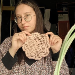 May include: A peach-colored, crocheted doily with a scalloped edge. The handmade piece features an openwork design with a floral pattern in the center. The doily is held up in front of a person.