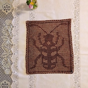 May include: A brown, crocheted doily with a detailed insect design. The doily is square and sits on a white tablecloth with a lace border. A small glass vase with colorful flowers is in the background.
