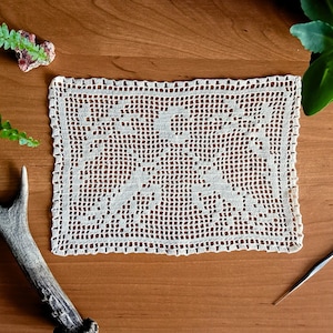 May include: Cream-colored, rectangular crocheted doily with a floral pattern. The doily is on a wooden surface, with a crochet hook, a deer antler, and greenery. The doily is a handmade item.