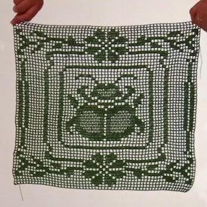 May include: A square dark green crochet doily featuring a central beetle motif surrounded by a geometric grid border and floral accents at the top and bottom edges. The intricate needlework creates a repeating pattern of open squares and solid stitches.