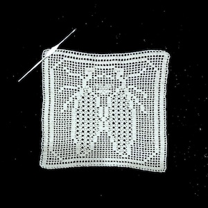 Op de afbeelding: Een vierkante, witte gehaakte doily met een gedetailleerd openwerkpatroon. De doily is geplaatst op een zwarte achtergrond, met een witte haaknaald op de hoek. Het ingewikkelde ontwerp heeft geometrische vormen en een decoratieve rand.