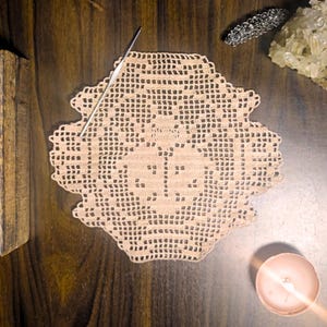 Puede incluir: Un tapete beige tejido a ganchillo con un patrón geométrico detallado, un ganchillo plateado, una vela pequeña, un grupo de cristales y una pulsera plateada están dispuestos sobre una superficie de madera oscura. El tapete es el foco central, mostrando intrincados trabajos de aguja.