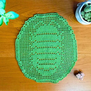 Puede incluir: Un tapete de ganchillo verde brillante con un borde festoneado. La pieza hecha a mano presenta un diseño calado con un patrón central. El tapete se muestra sobre una superficie de madera, junto a una pequeña suculenta en una maceta blanca.