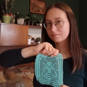 May include: A square, turquoise crochet doily with a floral design. The doily is held up in front of a person. The doily has a detailed openwork pattern and a scalloped edge. The background includes a wooden cabinet and various plants.