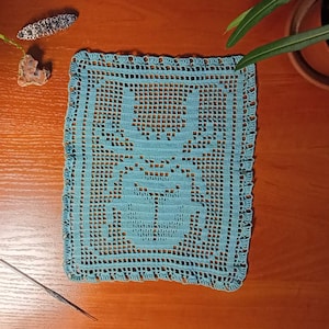 May include: A handmade, square, light blue crochet doily with a central floral design. The doily is placed on a wooden surface, with a crochet hook and decorative items nearby. The edges of the doily have a scalloped trim.
