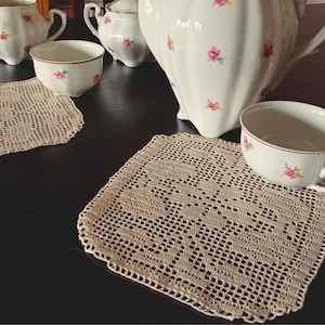 Może przedstawiać: Biały porcelanowy zestaw do herbaty z różowymi i niebieskimi kwiatowymi akcentami. Zestaw zawiera imbryk, cukiernicę, mlecznik i filiżanki. Widoczne są również dwie beżowe serwetki szydełkowe, dodające uroku vintage.