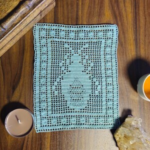 May include: A square, light blue crocheted doily with a central design, possibly a pineapple, sits on a wooden surface. A teacup with tea, a candle, a wooden box, and a crystal are also on the table.