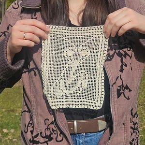 May include: Cream-colored, square doily with a floral design, held up against a person's clothing. The doily is made of crocheted thread, with a detailed openwork pattern. The background includes a brown belt and a mauve jacket.