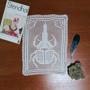 May include: A rectangular white crochet doily featuring a central beetle motif with intricate openwork patterns. The doily is displayed alongside a book titled Stendhal Czerwone i Czarne, a single dark feather, and a cluster of raw green crystals.