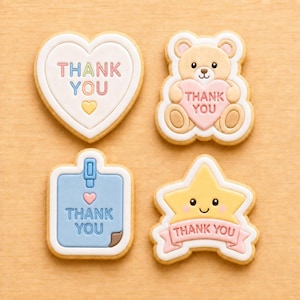May include: Four decorated sugar cookies with icing. One is a white heart with 'THANK YOU' in pastel letters. One is a tan teddy bear holding a pink heart with 'THANK YOU'. One is a blue square tag with 'THANK YOU'. One is a yellow star with 'THANK YOU'.