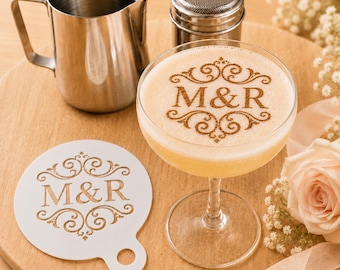 May include: A stainless steel milk pitcher, a metal shaker, and a cocktail in a glass featuring a brown cinnamon-dusted M&R monogram design. Beside the glass lies a white circular stencil with the same M&R monogram cutout design.