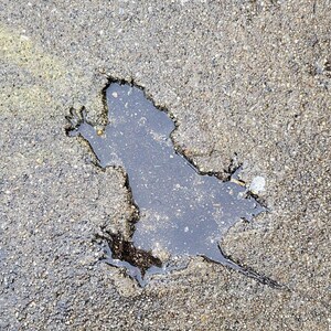 May include: A close-up of a puddle of water in a crack in the asphalt. The water reflects the sky and creates a silhouette of a frog.