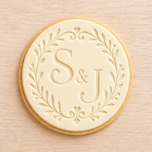 Peut inclure: Un biscuit rond recouvert d'un disque de pâte à sucre blanc cassé. Le centre présente les lettres S et J en relief, séparées par une esperluette, entourées d'une couronne de laurier délicate et symétrique avec de petits motifs de cœur.