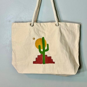 Puede incluir: Una bolsa de lona beige con asas de cuerda. La bolsa presenta un diseño pintado a mano de un cactus verde, un sol amarillo y una estrella dorada sobre un fondo rojo.