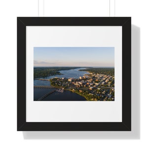May include: An aerial view of a city with a river winding through it. The city is surrounded by trees and water. The sky is blue and the sun is shining.