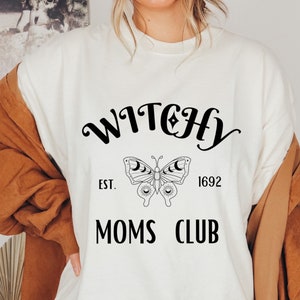 May include: Cream-colored t-shirt featuring the words "WITCHY MOMS CLUB" in black script. A butterfly graphic with moon and star details is centered below the word "WITCHY". The year "1692" is printed below the butterfly graphic.
