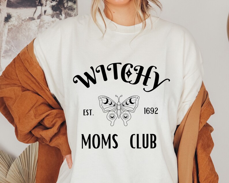 May include: Cream-colored t-shirt featuring the words "WITCHY MOMS CLUB" in black script. A butterfly graphic with moon and star details is centered below the word "WITCHY". The year "1692" is printed below the butterfly graphic.