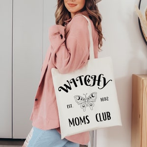 May include: Cream-colored tote bag with the words "WITCHY MOMS CLUB" in black script. A butterfly with moon details is centered below the word "WITCHY". The text "EST. 1692" is also included. The bag has a simple, minimalist design.