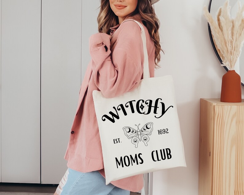 May include: Cream-colored tote bag with the words "WITCHY MOMS CLUB" in black script. A butterfly with moon details is centered below the word "WITCHY". The text "EST. 1692" is also included. The bag has a simple, minimalist design.