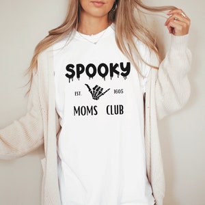 Op de afbeelding: Wit T-shirt met de zwarte tekst "SPOOKY MOMS CLUB", met een druipend lettertype en een skelet hand. Het shirt bevat ook de tekst "EST. 1605".