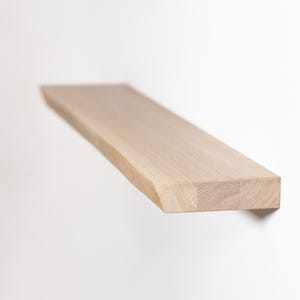 Puede incluir: Una estantería rectangular de madera clara, montada en una pared blanca. La estantería mide aproximadamente 76 cm de largo y tiene un acabado natural de vetas de madera. La estantería está diseñada para montarse en la pared para almacenamiento o exhibición.