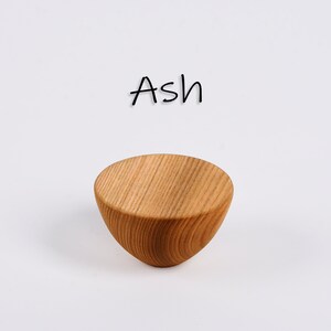 May include: A small, light brown wooden bowl with a rounded bottom and a flat top. The word "Ash" is written in black cursive above the bowl. The bowl has visible wood grain.