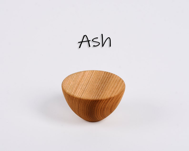 May include: A small, light brown wooden bowl with a rounded bottom and a flat top. The word "Ash" is written in black cursive above the bowl. The bowl has visible wood grain.