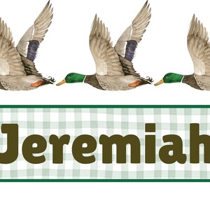 May include: Watercolor illustration featuring three flying ducks with outstretched wings. The ducks have green heads, brown bodies, and gray wings. Below, a green and white plaid banner displays the name "Jeremiah" in brown letters.