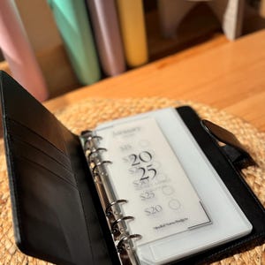 May include: A black leather bound planner with a clear plastic insert for a monthly budget sheet. The sheet has a title "January" and lists amounts in dollars for different categories.