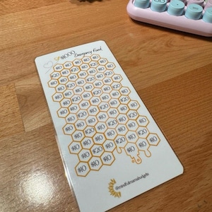 May include: A white card with a honeycomb design for tracking savings towards a $1000 emergency fund. The card features a series of hexagonal cells, each labeled with amounts of $10 or $20. The text "$1000 Emergency Fund" is at the top.