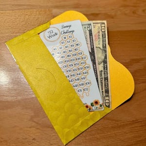 May include: A yellow envelope with a 52-week savings challenge card and U.S. currency. The card features a honeycomb design with amounts from $1 to $52. The envelope is open, revealing the card and several ten-dollar bills.