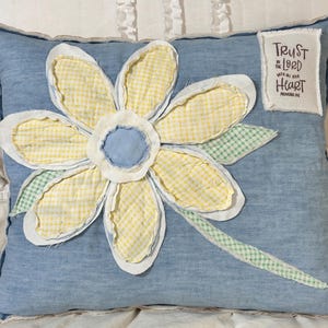 May include: A square blue denim throw pillow featuring a large fabric flower appliqué with yellow gingham petals, a light blue center, and green gingham leaves. A white fabric patch in the corner displays the text Trust in the Lord with all your heart.