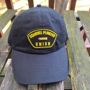 May include: Black baseball cap with a yellow patch that reads "Squirrel Plinkers Union" and a small image of a bullet.