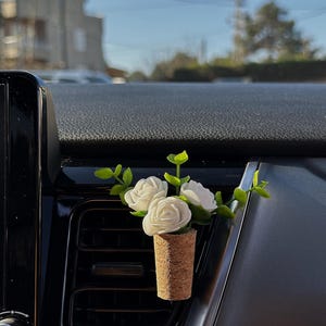 May include: A car air freshener vent clip featuring a natural cork base shaped like a wine stopper, topped with three small white artificial roses and green plastic leaves, securely attached to a black plastic vehicle air conditioning vent.