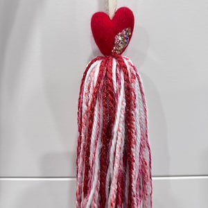 May include: A red felt heart with glitter accents attached to a white and red yarn tassel.