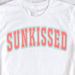 May include: White t-shirt with the word "SUNKISSED" in pink and green lettering. The shirt is laid flat on a white wooden surface with a pair of brown flip-flops, a pair of blue denim shorts, and brown sunglasses.