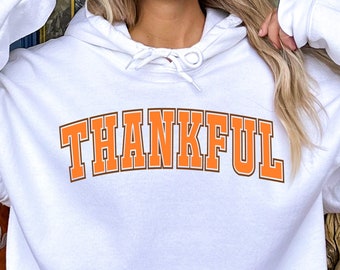 May include: White hoodie with the word "THANKFUL" in orange block letters. The hoodie has a drawstring hood and a front pocket. The image is set against a fall-themed background.