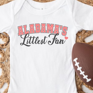 Puede incluir: Un body blanco para bebé con el texto "ALABAMA'S Littlest Fan" en letras rojas y negras con un pequeño corazón rojo. Un balón de fútbol marrón está acostado junto al body.