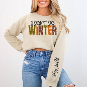 以下が含まれることがあります： ベージュのトレーナーに「I don't do winter」という文字が茶色、オレンジ、緑の文字で書かれています。右袖には「Very Well」という文字が黒い筆記体で書かれています。