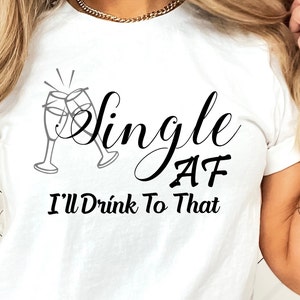 May include: White t-shirt with black text that reads "Single AF I'll Drink To That". The text is in a cursive font. There is a graphic of two champagne glasses clinking together above the text.