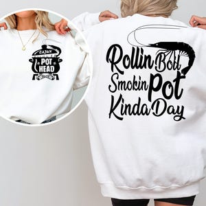 Puede incluir: Sudadera blanca con texto negro que dice "Rollin Boil Smokin Pot Kinda Day" y un gráfico de camarón en la espalda. La parte delantera de la sudadera tiene un gráfico negro de una olla con el texto "Cajun Pot Head".