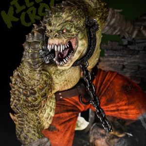 May include: A detailed sculpture of Killer Croc, a reptilian humanoid with green and yellow skin, sharp teeth, and red eyes. He wears an orange outfit and a metal collar with a chain. The text "KILLER CROC" is above the figure.