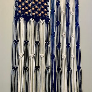Puede incluir: Un tapiz de macramé de pared negro, blanco y azul con cuentas de madera. El tapiz de pared tiene la forma de una bandera estadounidense.