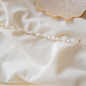 May include: A delicate pearl bracelet with a gold chain clasp. The bracelet is laid out on a white fabric background.