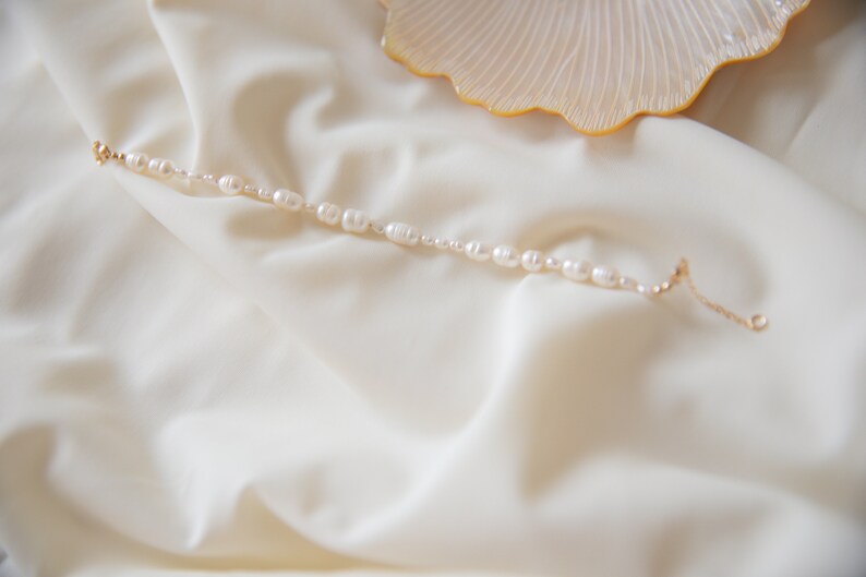 May include: A delicate pearl bracelet with a gold chain clasp. The bracelet is laid out on a white fabric background.